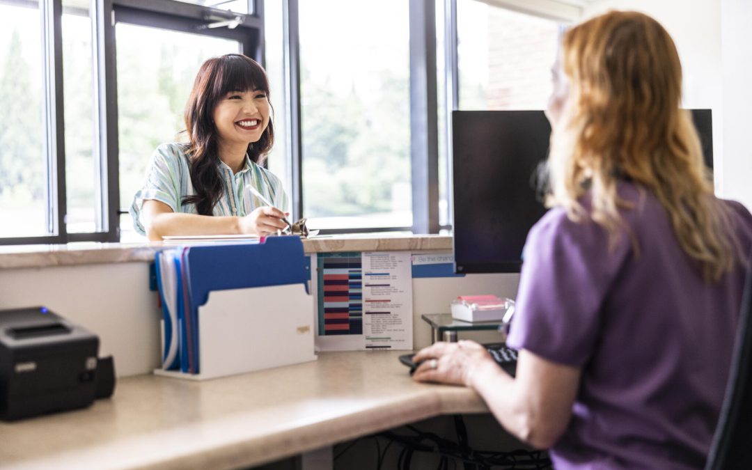 A smiling patient talking with a healthcare worker at a clinic front desk, representing how health insurance for small business employees supports well-being and culture.