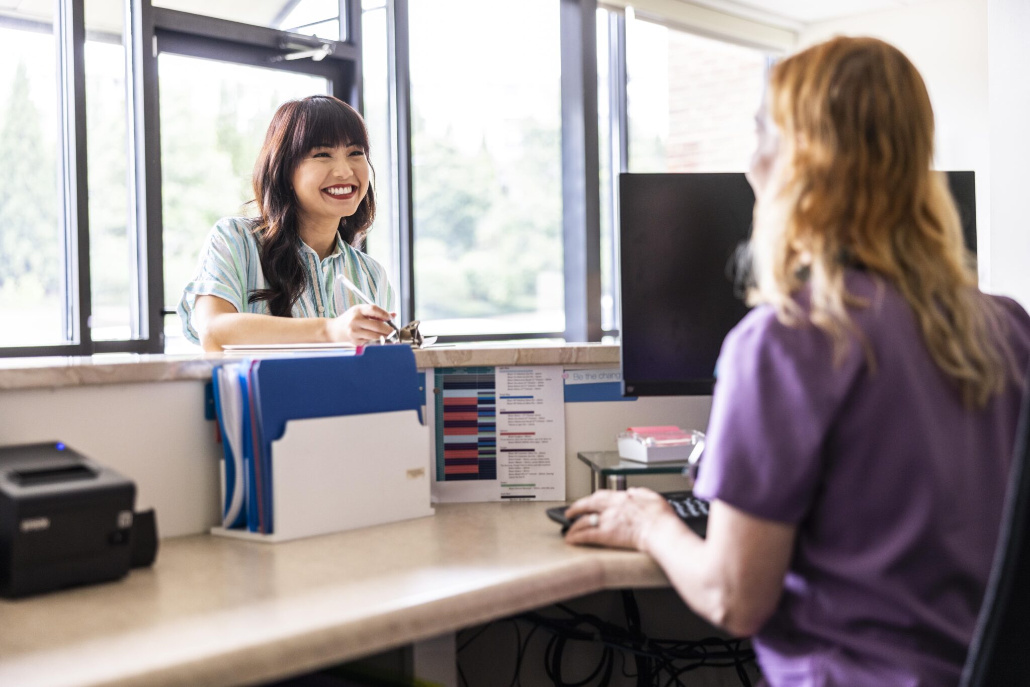 A smiling patient talking with a healthcare worker at a clinic front desk, representing how health insurance for small business employees supports well-being and culture.